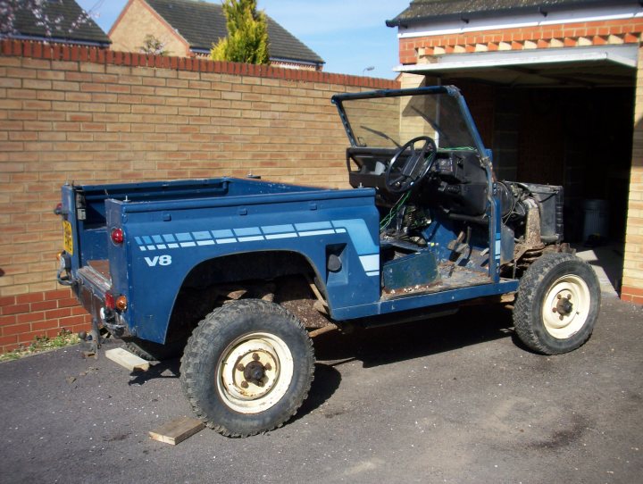 Defender V8 project - Page 1 - Readers' Cars - PistonHeads - The image depicts an old-style blue land rover parked on a street. The vehicle's cabin is open, revealing an interior that shows some wear and tear; the steering wheel is on the right side, indicative of the vehicle's possible origin as a left-hand drive vehicle. The land rover is parked in front of a building with a red brick exterior, partially obscured by a brick wall partially behind the vehicle. The context suggests a suburban or urban setting, possibly in the UK due to the style of the land rover.