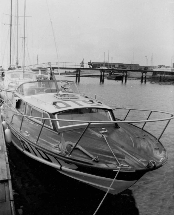 My Huntsman 31. - Page 3 - Boats, Planes & Trains - PistonHeads - The image is a black and white photograph capturing a serene maritime scene. Dominating the frame is a white sailboat with "S1" written on its transom. The boat is docked at a pier, surrounded by calm water. The pier leads to a bridge in the background where a train is crossing. Another boat can be spotted further down the pier, adding depth to the scene. The photo has an antique quality to it, suggesting it might be from the past.