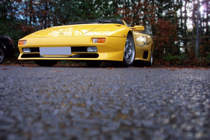 Lancashire Sunday Breakfast Club - Monthly Meet - Page 37 - North West - PistonHeads - The image depicts a vibrant yellow sports car parked on what appears to be a concrete surface. The car is situated facing the viewer, with its headlights on and a small amount of something red visible in the background. The car's design is sleek, featuring a prominent rear spoiler and a low-slung stance. The lighting in the photograph highlights the car's color and details, creating a striking contrast between the car and its surroundings.