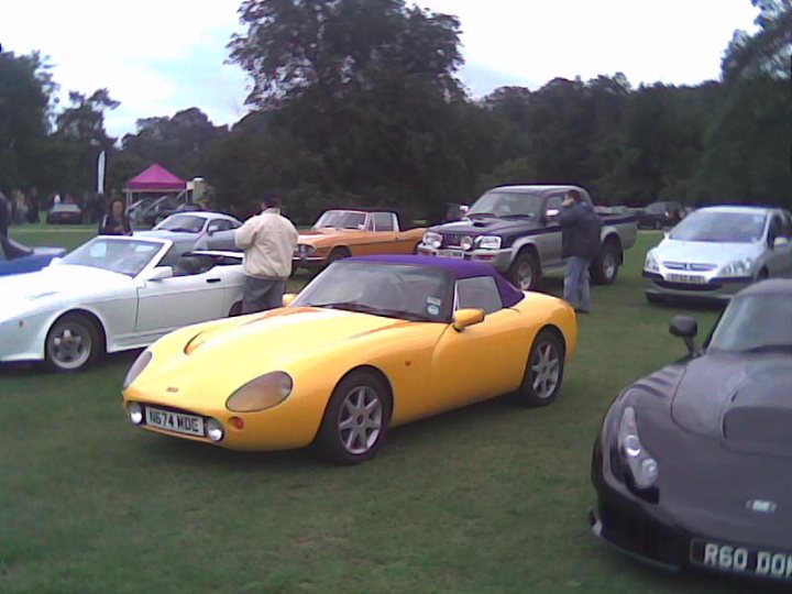 Proper Growl Pistonheads Ale - The image captures a lively outdoor event featuring a variety of vintage cars parked on grass. Dominating the scene is a vibrant yellow sports car, standing out due to its sleek and modern design. It's positioned near a white classic car, which adds a contrasting element to the scene. Further back, an orange classic car is parked alongside others, adding more color diversity to the display. A few people are scattered around the area, possibly admiring or discussing the cars. The setting seems like an open field or park, giving the event a relaxed and informal atmosphere. The presence of these classic vehicles suggests a passion for automotive history and design.