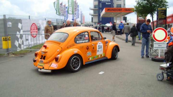 Weekend Pistonheads Eifelrennen - In the image, a distinctive scene unfolds on a street where a vintage Volkswagen Beetle is parked squarely in the middle. The car is painted in a vibrant shade of orange, drawing attention with its bright color. It displays the number "4200" prominently on its door and "504" on its side, suggesting it might be part of a car club or participating in a rally. The number "24" adorns the front license plate. In the background, people can be seen walking along the sidewalk, adding life to the scene. The cars and buildings in the vicinity hint at an urban setting.