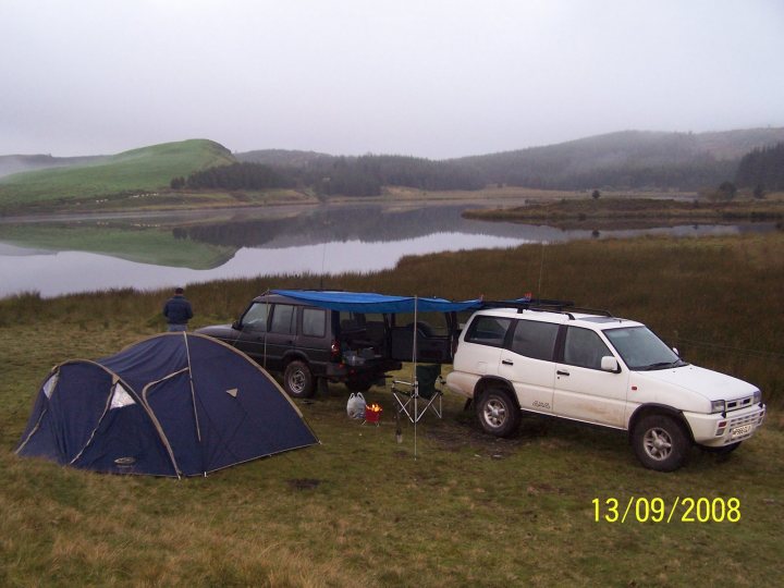 Anybody ever done an offroading / wild camping trip? - Page 1 - Off Road - PistonHeads - The image captures a serene camping scene in what appears to be a rural or natural setting. Two camping vehicles are parked next to each other on grass. A total of four people are present at the site, with one of them standing slightly ahead of the vehicles, facing towards them, and the other three located within the vehicles. The vehicles include two cars and a truck, and they are downcast on a grassy area by a river. The tent and chairs suggest a leisurely camping trip. The surroundings consist of grassy areas, a riverbank, trees, and hills, with a slightly overcast sky visible in the background.