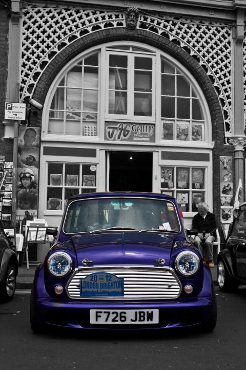 London to Brighton  - Page 1 - Classic Minis - PistonHeads - The image depicts a vintage-style garage with a curved front facade. A purple Mini Cooper car is parked in front of the garage with a clear glass windshield, the paint glistening under the light. The car bears a registration tag that reads "F726 JBW", and the grill indicates "LONDON BRIGHTON". In the background, there appears to be another car parked beyond the Mini Cooper, and to the right, there's a person sitting on a bench. The building has a neat, nostalgic appearance, with a visible lace-up attire detail. The overall setting evokes a sense of classic craftsmanship and automotive charm.