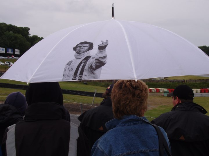 Coolest Umbrella EVER! - Page 1 - The Lounge - PistonHeads - This image depicts a scene at a racetrack. A group of people is seen standing under a large umbrella. On the umbrella is an image of the same man repeatedly stenciled on it, giving the impression that he is waving from the back. The racetrack is filled with a variety of cars and people scattered across the area. The umbrella primarily blocks out the rain, providing cover for the group underneath, while the image of the man adds a unique, humorous element to the scene.