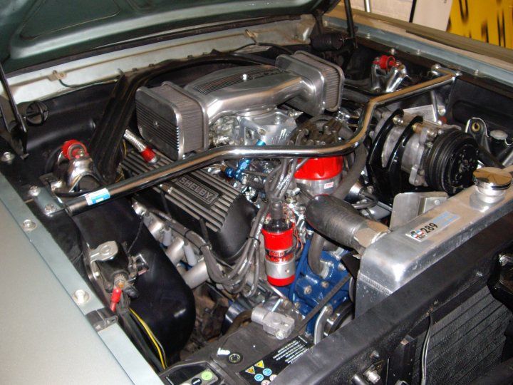 '65 mustang - Page 1 - Readers' Cars - PistonHeads - The image shows an engine compartment of a car, where the engine is exposed. The engine appears to be a V8 motor, with various components visible, including a large alternator, a battery in the back and multiple hoses and belts connecting various components. The engine bay is covered with a protective mat and the finish is a mix of polished metal and black atomized paint. The view is from the top looking down into the engine.