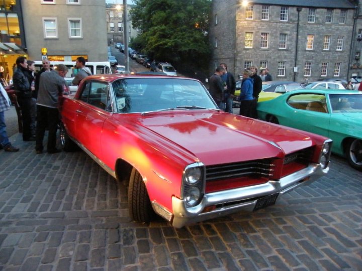 Edinburgh Cruisemeet Pistonheads - The image displays an outdoor scene with a focus on several vintage cars. A prominent red classic car is parked in the foreground on a cobblestone street. It features a reflective surface and silver rims. Standing near this car is a group of people, some engaged in conversation, who appear to be observing and possibly discussing the vehicles. In the background, to the right of the red car, several other vintage cars are parked, some appearing faded in comparison to the red one. The setting seems to be an urban area, possibly a street fair or similar event, given the presence of pedestrians and the casual atmosphere. The time appears to be dusk or evening, which may be evident from the lighting conditions and the portions of buildings and streetlights visible in the image.