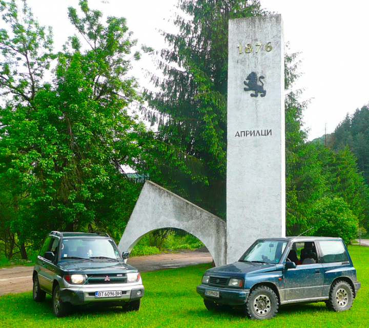 Little overnight trip to the Balkan range - Page 1 - Off Road - PistonHeads - The image captures a serene outdoor scene. Parked on the grass are two off-road cars, a gray jeep and a darker-colored SUV, partially obscured by the jeep's shadow. The vehicles are parked next to a stone monument, which is inscribed with Cyrillic text reading "1876" and a lion statue above it. This setting appears to be in the late afternoon or early evening, as evidenced by the soft light and the silhouettes of the trees in the background.