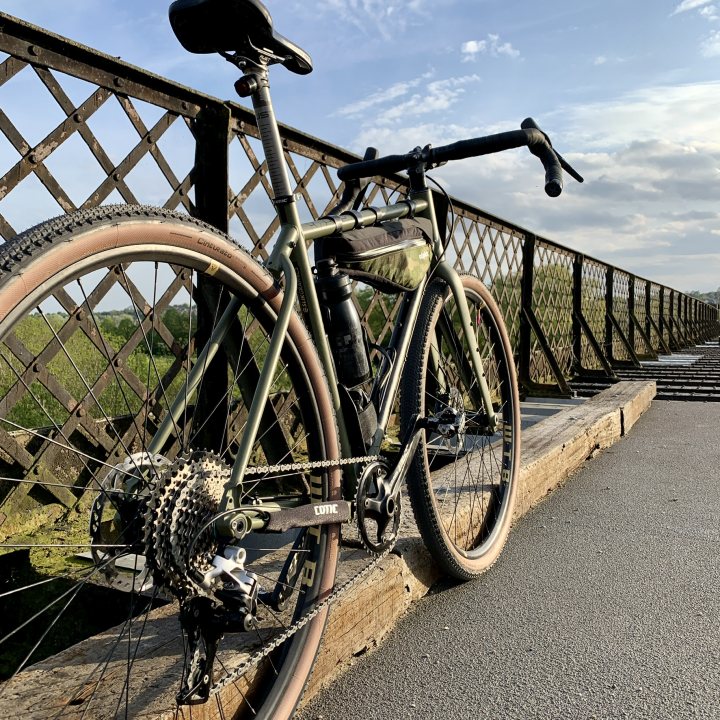 The "Show off your bike" thread! (Vol 2) - Page 88 - Pedal Powered - PistonHeads UK - The image shows a mountain bike leaning against a wooden railing on the side of a road. The bicycle is equipped with multiple gears, suggesting it's designed for off-road use or long rides. It appears to be parked in a rural setting, with a bridge and trees visible in the background. There is no text present in the image.