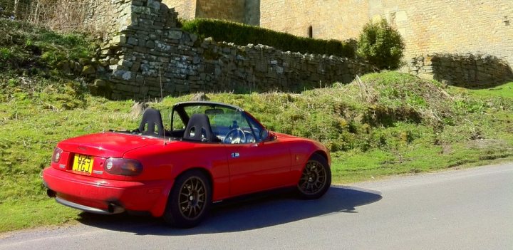 Lane Pistonheads Conundrum Rollbar - The image captures a vibrant scene of a red sports car parked on a grassy section of a road next to a stone wall. The car, a convertible, is positioned in the foreground, its top down, showcasing two black seats and a yellow license plate. The road itself is lined with a stone wall on one side and a lush green hillside on the other, adding a sense of tranquility to the otherwise dynamic image.