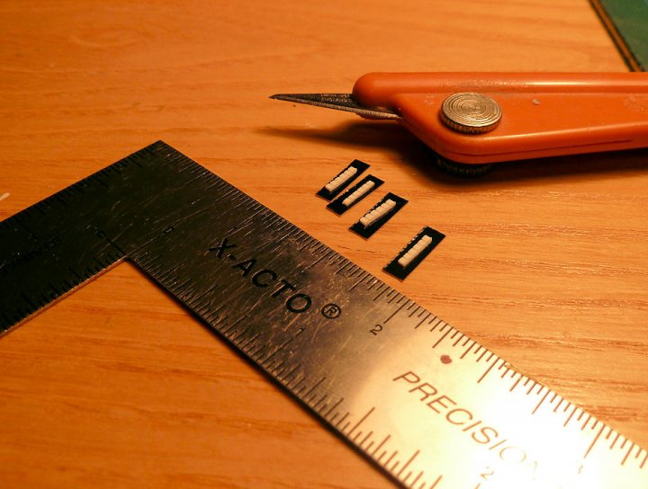 A pair of scissors sitting on top of a table - Pistonheads - The image shows a wooden surface featuring a metal ruler, a pair of scissors, and some stretches of what appear to be black elastic or rubber. The ruler has black measurements and the brand name "xacto" in white letters, indicating it's a precision ruler, likely frequent across offices and schools. The PRECISION label is in a white rectangle on a black background, a classic design choice presumably to enhance visibility and precision. The stretches of black material on the surface suggest a DIY project or art intent. The scissors are positioned beside the ruler, along with the elastic, possibly used in the current task.