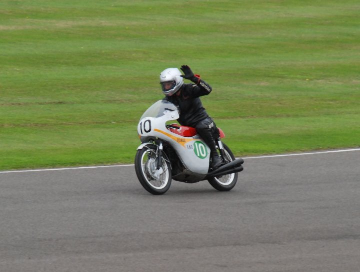 Sunday Bike Parade - Page 1 - Goodwood Events - PistonHeads - The image features a motorcyclist riding on a race track. The rider is wearing a red and black suit, a white helmet, and is greeting the audience with a raised hand. The motorcycle is white with red and black accents, and it also displays "10" prominently. The background shows a grassy area and part of the track, suggesting this might be a professional race.