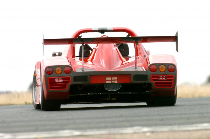 Trackday silencer - Page 1 - Radical - PistonHeads - The image depicts a bright red racing car with a large, aerodynamic front spoiler and rear wing, suggesting a high-performance design. The car is moving on a tarmac road, which has a thin white line indicating a lane boundary. The vehicle appears to be leaning slightly to the left, possibly due to its motion or while navigating a curve. The lighting suggests either a cloudy day or a location with diffuse lighting.