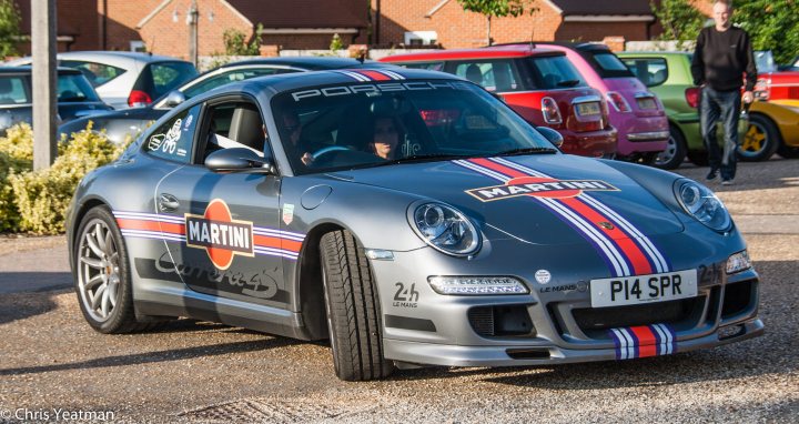 A car that is sitting in the street - Pistonheads - In the image, there's a dark grey sports car with a white stripe on its side. The license plate of the car is clearly visible. The car is parked in a lot filled with various other vehicles. There are two people in the image: one is walking towards the car and the other is behind the car. The background features a brick building, and there's a potted plant in the foreground. The lighting suggests it's either dawn or dusk, with the sky having a purple hue.