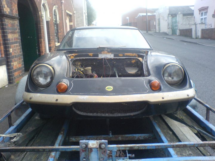 Jps Europa Pistonheads - The image shows an antique car being transported on a flatbed trailer. The exterior of the car is black with visible scratches and signs of wear. The hood is missing, exposing a partially intact engine. It appears the vehicle was involved in an accident but is now being moved away from the scene, possibly heading towards a repair shop. The setting suggests a rural environment with a brick or stone building and a typical street scene in the background.