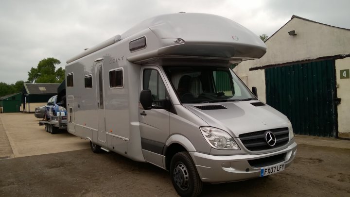 Show us your gear (tents to motorhomes) - Page 18 - Tents, Caravans & Motorhomes - PistonHeads - The image shows a gray trailer with a mercedes-benz logo, parked on a gravel lot. The trailer is equipped with a large, white overhang at the front, suggesting it might be used for horse or livestock transportation. In the background, you can see the outlines of a greenfield and a white building with a green door, partially obscured by the trailer. There are also a couple of vehicles and a horse trailer visible in the background, indicating a facility or storage area for vehicles. The sky is cloudy, giving the scene a neutral tone.