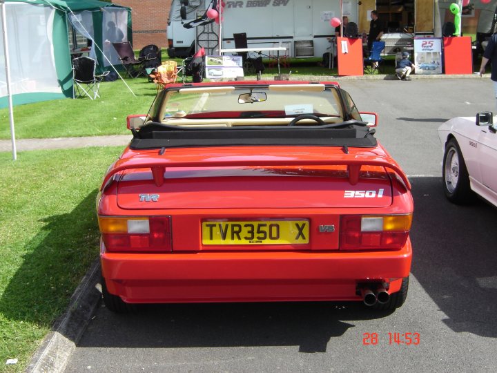 The Ultimate Christmas Present - Page 1 - Wedges - PistonHeads - The image captures a vibrant scene at a car show. A red TWR350 X, standing out prominently in its distinctive color, is parked on a shaded street with grass on the side and a white tent in the background. The car's registration number is clearly visible, adding an element of authenticity to the scene. The setting suggests a casual yet special event, possibly a gathering of enthusiasts or a promotional event for the TWR350 X model.