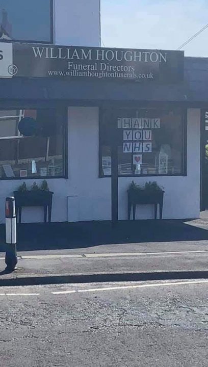 Coronavirus Humour - Page 25 - The Lounge - PistonHeads - The image shows a street scene with a prominent sign for "William Houghton Funeral Directors." In front of the store, there are two small plants. Adjacent to the funeral director's sign is another sign that reads "Hugh Jones M&S" in a slightly smaller font size. There is a window display on this building with some books and an item that resembles a vase or jar. A parked car can be seen across the street from these establishments. The sky above appears overcast, suggesting a cloudy day.