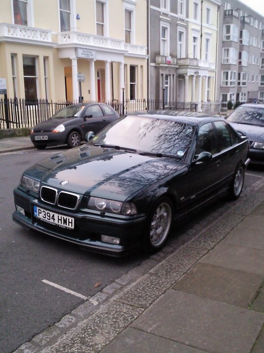 Best picture of your car (current or old) - Page 30 - General Gassing - PistonHeads - The image shows a scene from a city street where two cars are parked. The most prominent car is a dark-colored, small-sized sedan. This vehicle has a shiny exterior reflecting the overcast sky. In the background, partially obscured by the cars, are buildings indicative of a residential area. The overall atmosphere of the image is one of a quiet, suburban day with an overcast sky suggesting impending rain. The presence of license plates suggests the location is within the United Kingdom.