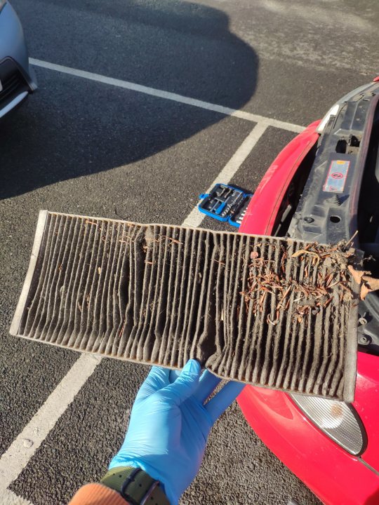 B5 Audi A4 2.8 Quattro Avant - Page 3 - Readers' Cars - PistonHeads UK - The image is a photograph featuring an automobile air filter that is dirty and needs replacement. A hand holding the air filter is visible, suggesting that the photo was taken by someone inspecting or cleaning the car's engine bay. The background shows a partially obscured vehicle with a red door visible, indicating that the focus of the image is on the air filter. There are no visible texts or logos in the image.