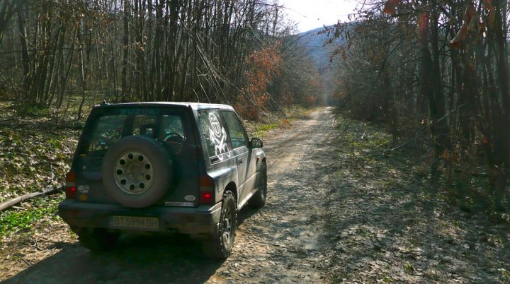 Winters over... Big chunky proper tyres back on... - Page 1 - Off Road - PistonHeads - The image is a photograph of a scene from nature. The most prominent object in the scene is an old, dark gray four-door SUV parked on a muddy road. This road is surrounded by lush greenery and is bordered by dense forest or woodland on both sides. The foliage is large and seems to suggest that the image was taken in autumn, as the leaves of the trees are a mix of green and autumnal shades of red and yellow. The sky is overcast, and it seems to be a cloudy day. There are no visible texts or distinctive markings on the SUV or in the immediate surroundings that would indicate a specific brand or model. The scenery appears relatively undisturbed and peaceful.