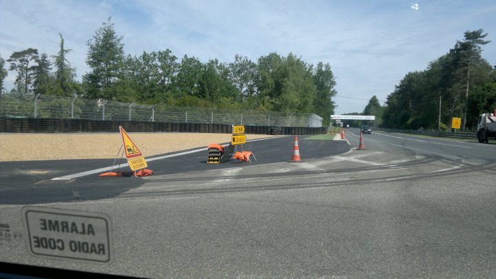 Arnage corner modifications  - Page 1 - Le Mans - PistonHeads - The image depicts a section of a road adjacent to a line of orange traffic cones. There are four orange cones evenly spaced along the length of the coned pass. The ground in front of the cones appears to be covered with loose gravel or dirt. A black guard rail with a series of slats runs for much of the length of the scene, providing some separation between the road and the construction or maintenance area. The surrounding environment suggests a rural or suburban area with trees and other vegetation in the background. The atmosphere is overcast with a gray and cool sky.