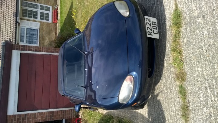 A man is standing next to a motorcycle - Pistonheads - This image shows a dark blue sports car parked on a driveway. The car's sleek design and glossy finish suggest it is newer or well-maintained. The number plate on the front of the car reads "R22COB," indicating a licence from the UK. The driveway appears to be made of concrete and is lined by a rustic brick wall and a grassy area with a few scattered flowers. The background consists of a home with a distinctive shaped window.