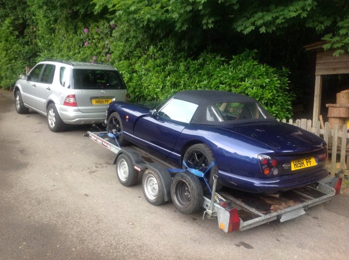 Random TVR pics - Page 409 - General TVR Stuff & Gossip - PistonHeads - The image shows two cars parked on a driveway. On the left, there's a light gray sedan, while on the right, a blue sports car with a black and white top is visible. The sports car is being towed by a trailer that is attached to a separate vehicle. The trailer has four wheels and appears sturdy enough to handle the weight of the sports car. The background features green foliage and a wood structure, suggesting a residential area.