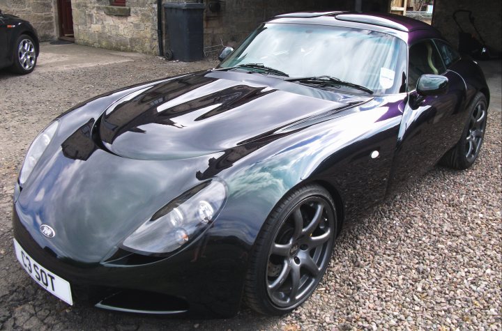 Ill Start Pistonheads - The image showcases a black convertible car with a distinguishing flared top design, exhibiting a shiny surface reflecting the surroundings. A person is partially visible at the edge of the frame, yet their role or activity is unclear. A car wash vacuum cleaner can be seen on the side, suggesting that the car has been recently cleaned or is awaiting cleaning. The setting appears to be a gravel-covered ground with a stone building or wall in the background, possibly a backdrop for the car's storage or display.