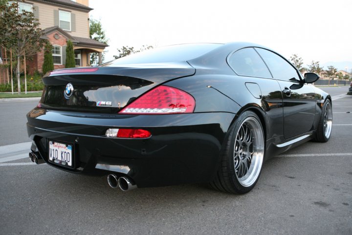 Pistonheads Decently Modified - In the image, a sleek black BMW is parked in a lot, facing the viewer. The vehicle is a two-door model, with visible modifications such as a rear spoiler, sporty side skirts, and a fitted lip bumping the front lip of the car. The BMW logo is prominently displayed on the trunk. The car is parked near a sidewalk with a lamppost, and there are buildings in the background. The setting suggests a residential area or possibly a business district with a lot designated for parking.