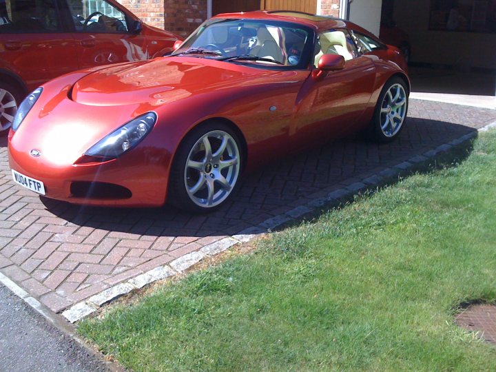 Pistonheads - This is an image of a red sports car parked on a brick patio. The car features a sleek design with an open top and large, round alloy wheels. Its surface reflects the sunlight, enhancing its red color. Behind the car, there are other, less prominent vehicles in a garage, and beyond them, a lawn leading to the garage. There's a slight reflection of the scene on the brick paving, adding depth to the picture.