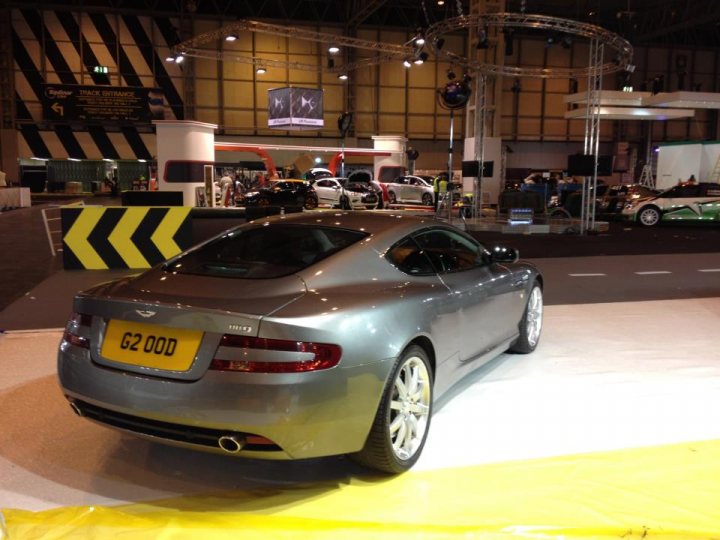 Top Gear Live (NEC this weekend) - Page 1 - Aston Martin - PistonHeads - The image features a silver sports car parked on a showroom floor. The car is positioned at a three-quarter angle, showcasing its sleek design and the license plate is clearly visible. The background of the photograph reveals a convention-like setting with bright lighting and other vehicles visible. A yellow caution pole is also noticeable in the background, giving the impression of a controlled environment.