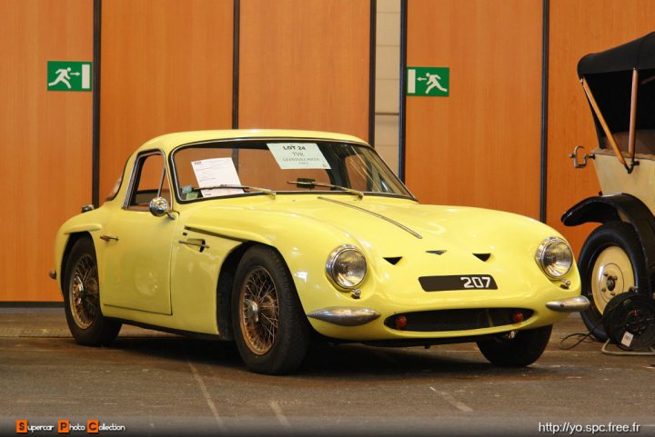 Early TVR Pictures - Page 4 - Classics - PistonHeads - The image features a beautiful antique car parked inside a building, possibly a museum, given the vintage style of the car and the architecture of the setting. The car is yellow, with chrome details that gleam under the indoor lighting. It appears to be a convertible with black text on the side, providing contrast against the car's yellow exterior. To the right of the car, there's a handwritten sign leaning against a yellow and black panel, but the text is too small to be legible. The overall scene suggests an exquisite collection on display.