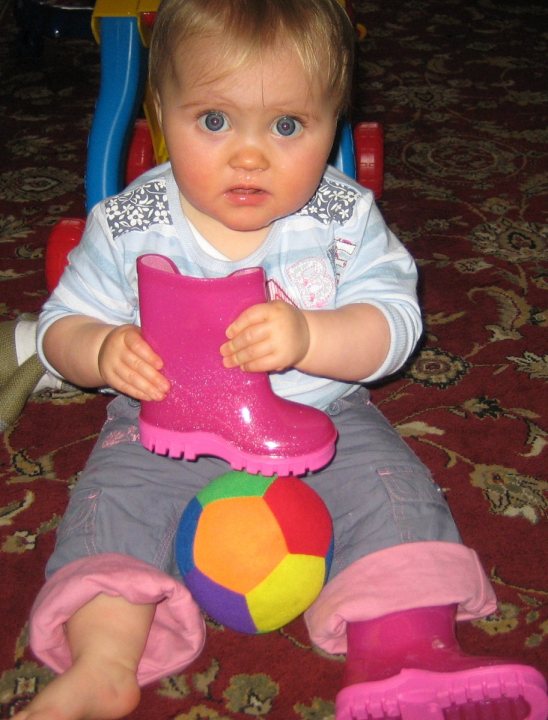 In the image, a little girl is seen sitting on the floor with a striking gaze emerging from her large blue eyes. She is holding a toy, specifically a pair of pink rain boots. A multicolored, inflatable ball lies next to her on the floor. The setting appears to be a brightly colored indoor room with a carpeted floor and various toys scattered around. The girl's vibrant green shirt contrasts with the predominant colors of red and yellow in the room.