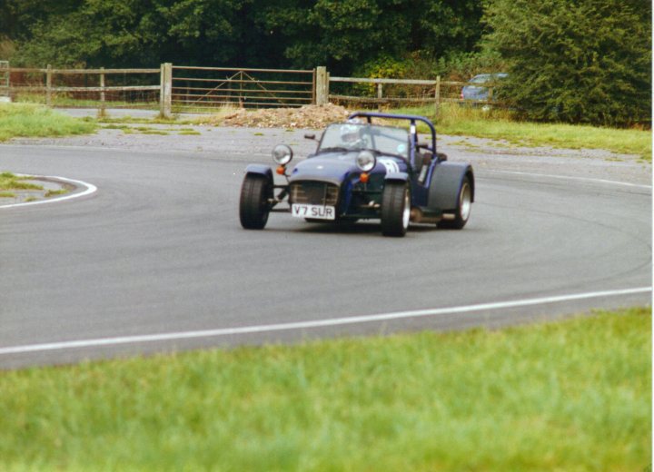 Pistonheads Games Oppo Dap - This image captures a dynamic scene of a vintage vehicle driving on a winding road. The vehicle appears to be a small convertible with a round top, possibly a Morgan sports car. It's traveling around a curve with a backdrop of lush greenery, including trees and a field, suggesting a rural or semi-rural setting. The road has clear lane markings indicating a well-maintained public road. The car's number "V7 SUC" is visible, adding a sense of personality or brand association to the scene. The image conveys a sense of motion and the timeless appeal of classic automobiles.