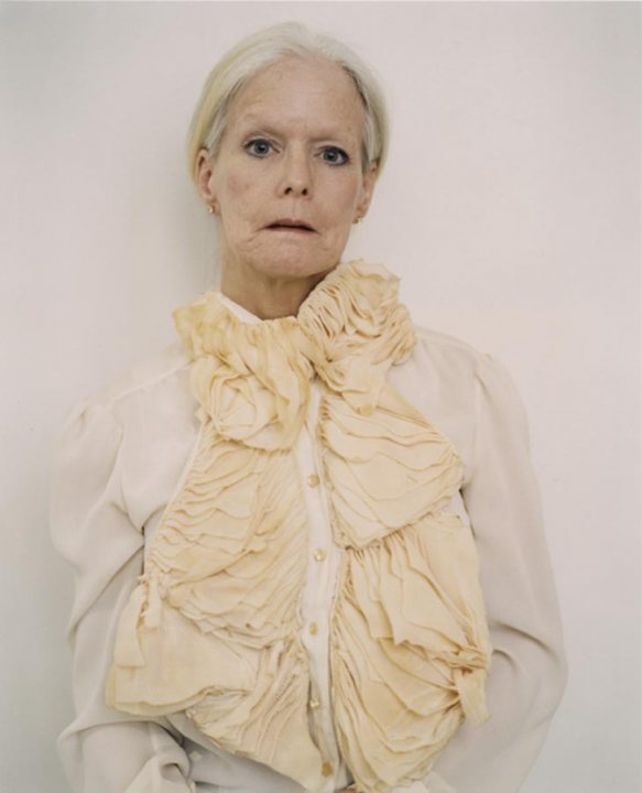 The image is a photograph of a person. The person appears to be an elderly individual with light-colored hair and fair skin, and they are wearing a beige garment with a frilly collar that has a textured fabric. The person's expression is neutral, and their gaze seems directed towards the camera. The background of the image is solid and light-colored, providing a plain backdrop that contrasts with the subject's attire. There are no distinct features, logos, or texts visible in the image.