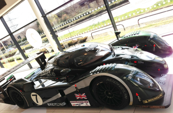 Bentley Lap of Honour Tour - pics from Bentley Manchester - Page 1 - Rolls Royce & Bentley - PistonHeads - The image shows a sleek, modern-looking race car indoors, where its polished surface reflects light. The vehicle has a predominantly black and white color theme with British racing flag accents. To the right of the car, there is a glimpse of another similar vehicle, suggesting that this might be a showroom or exhibition setting. A building with glass windows is visible in the background, providing ample natural light that brightens the scene. There are no visible texts or distinctive features in the image that provide additional information about the make or model of the car.