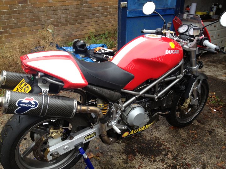 A motorcycle parked on the side of a road - Pistonheads - The image showcases a Ducati motorcycle parked on a street. The motorcycle is a striking combination of red and black with gold accents. It's a sportbike model with a lit-up "Ducati" emblem on its side. The motorcycle is leaning against a wall on its kickstand.