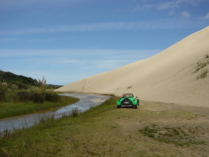 Pistonheads Beach Mile - In the image, a lone green vehicle is driving on a sandy road that splits into two, like a fork. The scene takes place in a beige desert, which is contrasted by the green vegetation to the left of the road. A blue stream of water is also visible in the background, adding a splash of color to the otherwise monochrome landscape. The perspective of the photo is from above, providing a bird's eye view of the vehicle, the road, and the surrounding environment.