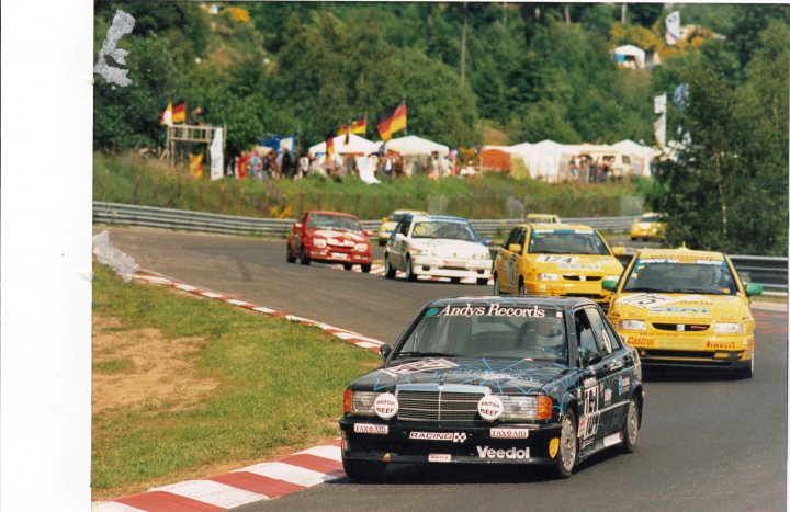 Pistonheads - The image appears to be a photograph capturing a moment on a racing track during a car event. The track is wide and curves gently to the right, with a barrier visible on the left side. Various racing cars are present in the scene, each displaying different sponsor logos and vibrant colors. The one closest to the viewer is black, red, and white. In the background, there is a crowd of spectators, and the landscape is framed by mature trees. The sky is partly cloudy, suggesting an outdoor daytime setting.