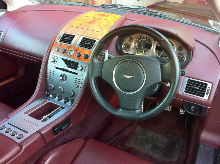 My First Aston - Page 1 - Aston Martin - PistonHeads - The image shows the interior of a car, featuring the driver's side of the cockpit area. At the forefront is the steering wheel, which is black and has the Mercedes-Benz logo in the center. The car seems to be a convertible, as the top is not visible. The car has a black interior with red trim, particularly noticeable on the driver's door side, which has matching red leather handle inserts. The dashboard also has a wooden panel with red trim that frames the control buttons and air vents. The vehicle appears to be well-maintained, indicative of a luxury brand.