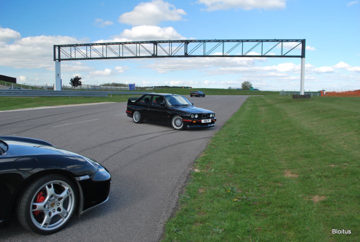 Pistonheads Service Supply Silverstone Snaps Sunday - The image depicts an open road with a black, sports-style vehicle curving around the right side of the frame. In the background, there is a suspension track with barriers and a sign above it, and another black car is parked parallel to the track, possibly behind a guard rail. The sky is clear with a blue hue, and the lighting suggests it might be late afternoon or evening. The setting appears to be a public road or racetrack with visible run-off areas and safety measures.