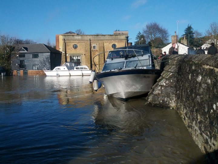 Maidstone town centre flooded - Page 1 - Kent & Essex - PistonHeads - The image depicts a serene scene with a small boat gently crashing into a sturdy stone wall near a body of water. In the background, two buildings can be seen with a striking contrast: one is a modern, gray structure with white top, and the other is a traditional brick house that adds charm to the setting. The docked boat has white both sides, and several people are visible in the vicinity, adding a sense of activity to the tranquil environment.