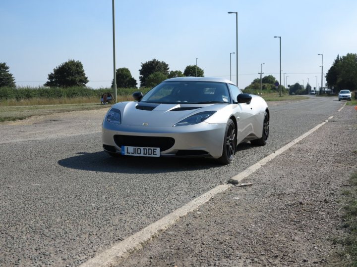 I've also bought one.... - Page 1 - Evora - PistonHeads - The image captures a dynamic scene on a road. Dominating the frame is a sleek silver sports car, its front facing towards the right side of the image. It's emblazoned with the license plate "LJIO DE", suggesting its origin or perhaps indicating a lease attachment. The car's metallic sheen stands out against the background of a clear sky. The vehicle is parked on the left side of the road, adjacent to the shoulder, with a grassy verge extending from the curb. Adjacent to the car is a lone telephone pole, adding an element of human-made infrastructure to the otherwise natural landscape. In the distance, other cars can be seen on the road, adding depth to the scene and providing a sense of scale. The car appears to be in good condition, ready to roar down the road.