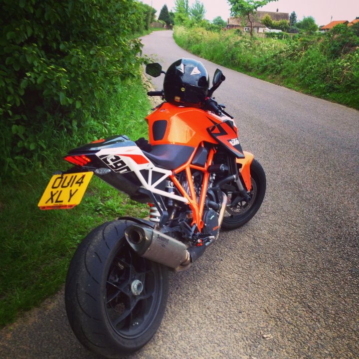 KTM 1290 SDR Suffolk today  - Page 1 - East Anglia - PistonHeads - The motorcycle in the image is a striking combination of orange and black with white accents. The bike is parked on the side of a road in a rural area, the lush greenery visible in the background suggesting a peaceful countryside setting. The bike's single rider's helmet is placed on the seat, indicating that the rider has momentarily stepped away from the motorcycle. The bike's dual exhaust pipes add to its sporty appearance. The number plate of the motorcycle, BL44 IXL, stands out prominently on the side of the bike.