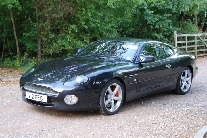 To those of us with 'older' Astons... - Page 1 - Aston Martin - PistonHeads - The image presents a sleek, black sports car parked on a gravel road, which cuts through a serene landscape of lush green trees and an expanse of grass. The car's license plate, reading "V12 PFG", is visible to the left of the front wheel. A wooden gate can be seen in the background, indicating a private or rural setting. The overall scene suggests a tranquil day, perhaps filled with the joy of driving, as evidenced by the sunlight shining on the car, partially visible due to a part of the frame being shielded from view.