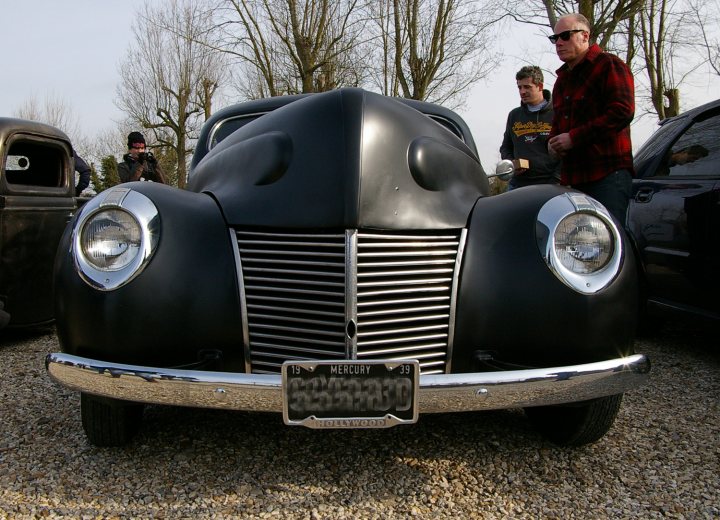 Goodwood Breakfast Club - Sushi Sunday - photos - Page 1 - Goodwood Events - PistonHeads - The image features a large, old-fashioned automobile painted in a bluish black color, reminiscent of a classic era. The vehicle has a distinctive grill pattern and chrome detailing, making it stand out. In the background, there are people present; the first appears to be a man in glasses standing beside the car while the second, possibly a woman in a red garment, is a bit further away. The scene seems to be in an outdoor setting, likely during the day under overcast or slightly cloudy sky. The vintage car, with its imposing size and classic design, is the most prominent object in the image, while the people are secondary, providing a sense of scale and human interest to the scene.