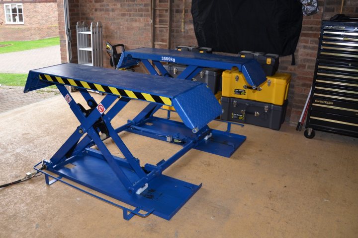 Pistonheads Buying Ramp Garage - This image shows a garage workspace with various equipment. In the center of the image is a sturdy blue piece of machinery, likely a hydraulic lift or a bench with hydraulic support. To the right, a stack of gray and yellow plastic storage containers is neatly arranged. On the left side, there is a yellow metal cabinet with a lock on it. The background features a brick wall, providing a contrast to the modern industrial equipment in the foreground.