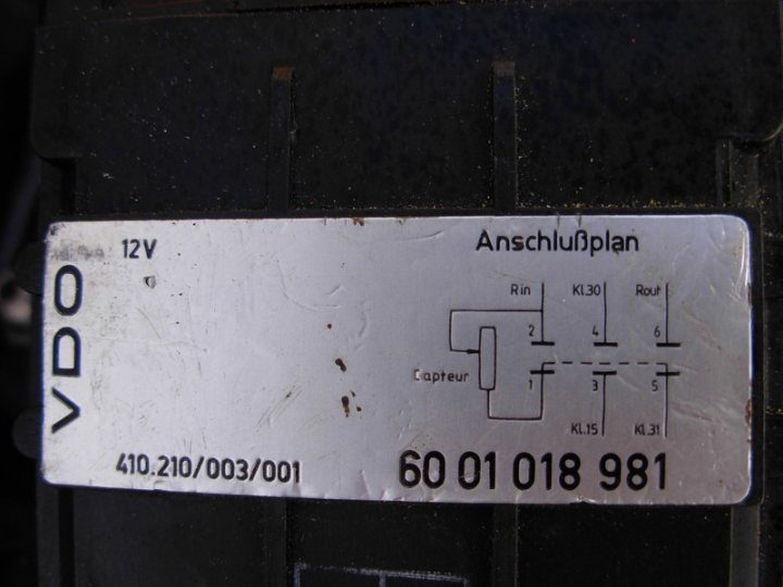 A sign that is on the side of a building - The image shows an identifier label on a mechanical object, presumably a part of an electrical or vehicle system. The label has a barcode, serial number, and important information such as "12V" which refers to a 12-volt unit, and the word "ANSCHLU", which means "connection" in German, indicating plugs or electrical ports. There is also a handwritten note above the barcode. The date and time written on the label are "410.210/03/01" and "60 01 018 981". The label's information appears to refer to a specific model or designation of a mechanical part.