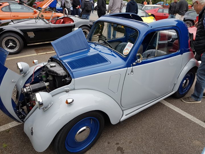 The Best ///M/Barge/General Rant/Look at this/O/T(Vol XVIII) - Page 179 - General Gassing - PistonHeads - The image features a classic, blue and silver, Volkswagen Beetle convertible car parked in a row of other vehicles in what appears to be an outdoor show car setting. The car's hood and battery are prominently visible, revealing its engine and battery components. On the car, there's a small sign that could indicate the car's model, make, or a customized message. In the background, several other people are standing, possibly attendees or individuals associated with the event or car owners.