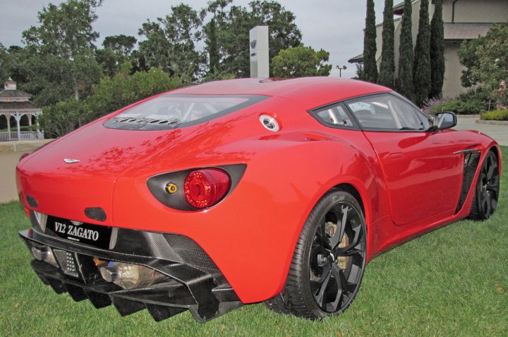 Pics from Aston event, Monterey - Page 1 - Aston Martin - PistonHeads - This image features a bright red sports car parked on a grassy area. The car is sleek and polished, with a curved silhouette typical of sports vehicles. The wheels are large with a multi-spoke design, enhancing the vehicle's aerodynamic appearance. There is a slight reflection in the ground behind the car, suggesting a wet or recently rained-upon surface. In the background, partially obscured, there are trees and a house, indicating that this scene is possibly in a residential area. The car has a textured rear wing and a rear bumper plate that displays "V12 ZAGATO," hinting at a luxury or high-performance model.